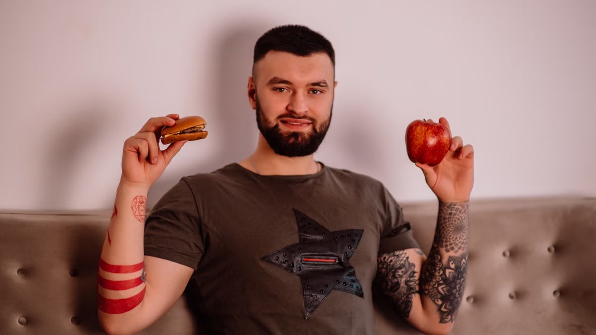 Bearded man pondering diet choice with apple and burger. Indoors with tattoos visible.